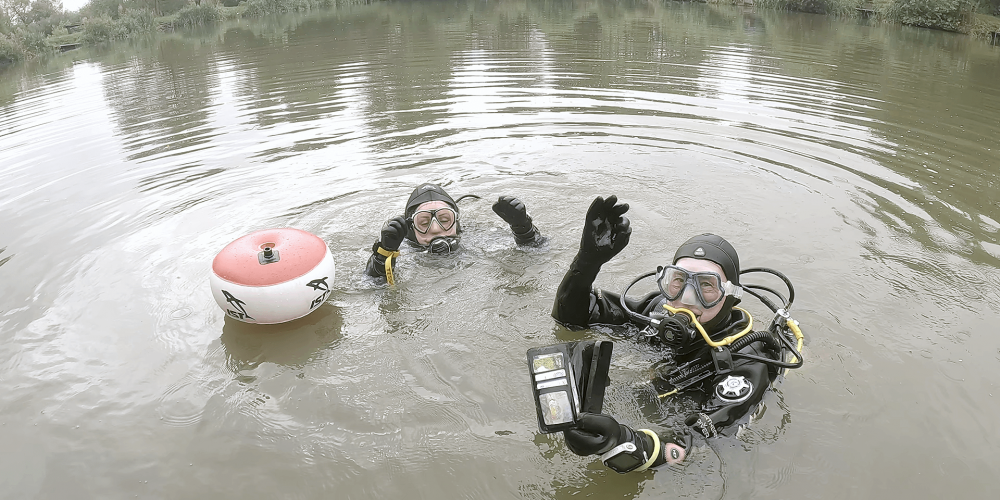 The image shows two of our divers, both smiling broadly with visible joy and excitement after successfully retrieving a lost phone from the lake.